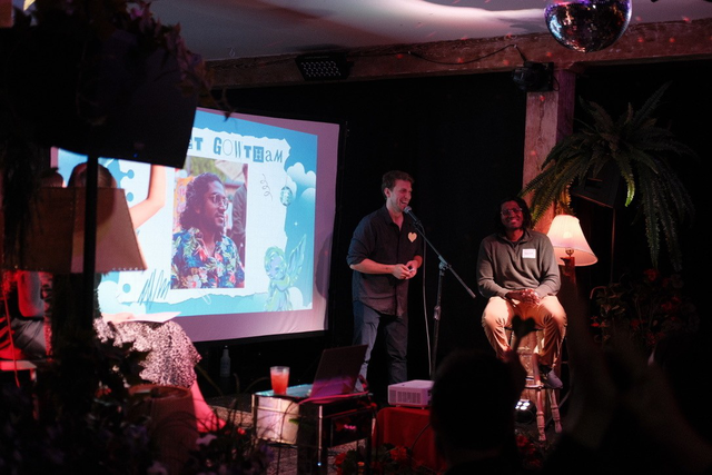 A man speaks into a microphone while another sits on a stool, with a projected slide featuring a photo and text behind them in a dimly lit room.
